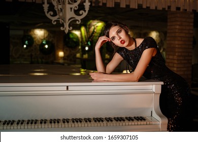 Portrait Of 20s Style Festive Beauty In A Restaurant. Young Beautiful Woman In Art-deco Style, In Black Dress In A Luxurious Interere Chicago 20s, The Era Of Gangsters. Singer  At The White Piano