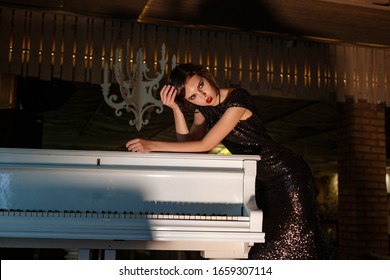 Portrait Of 20s Style Festive Beauty In A Restaurant. Young Beautiful Woman In Art-deco Style, In Black Dress In A Luxurious Interere Chicago 20s, The Era Of Gangsters. Singer  At The White Piano