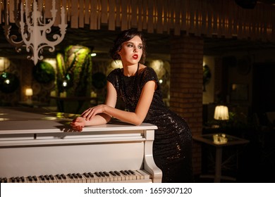 Portrait Of 20s Style Festive Beauty In A Restaurant. Young Beautiful Woman In Art-deco Style, In Black Dress In A Luxurious Interere Chicago 20s, The Era Of Gangsters. Singer  At The White Piano