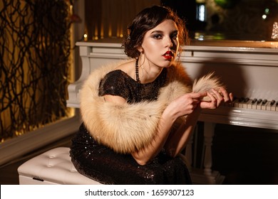 Portrait Of 20s Style Festive Beauty In A Restaurant. Young Beautiful Woman In Art-deco Style, In Black Dress In A Luxurious Interere Chicago 20s, The Era Of Gangsters. Singer  At The White Piano