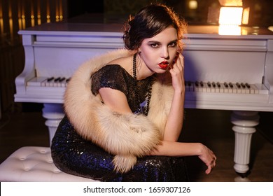 Portrait Of 20s Style Festive Beauty In A Restaurant. Young Beautiful Woman In Art-deco Style, In Black Dress In A Luxurious Interere Chicago 20s, The Era Of Gangsters. Singer  At The White Piano