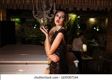 Portrait Of 20s Style Festive Beauty In A Restaurant. Young Beautiful Woman In Art-deco Style, In Black Dress In A Luxurious Interere Chicago 20s, The Era Of Gangsters. Singer  At The White Piano