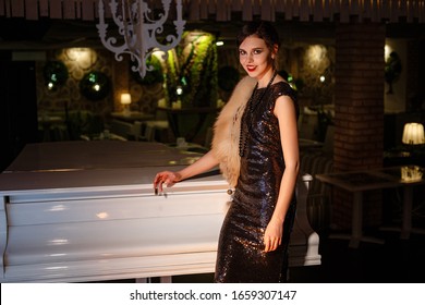 Portrait Of 20s Style Festive Beauty In A Restaurant. Young Beautiful Woman In Art-deco Style, In Black Dress In A Luxurious Interere Chicago 20s, The Era Of Gangsters. Singer  At The White Piano