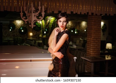 Portrait Of 20s Style Festive Beauty In A Restaurant. Young Beautiful Woman In Art-deco Style, In Black Dress In A Luxurious Interere Chicago 20s, The Era Of Gangsters. Singer  At The White Piano