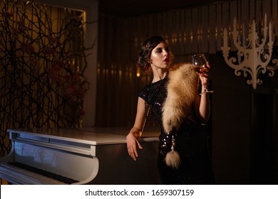 Portrait Of 20s Style Festive Beauty In A Restaurant. Young Beautiful Woman In Art-deco Style, In Black Dress In A Luxurious Interere Chicago 20s, The Era Of Gangsters. Singer  At The White Piano