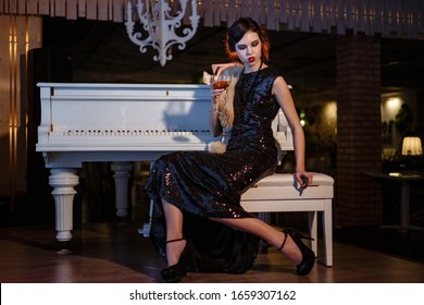 Portrait Of 20s Style Festive Beauty In A Restaurant. Young Beautiful Woman In Art-deco Style, In Black Dress In A Luxurious Interere Chicago 20s, The Era Of Gangsters. Singer  At The White Piano