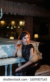 Portrait Of 20s Style Festive Beauty In A Restaurant. Young Beautiful Woman In Art-deco Style, In Black Dress In A Luxurious Interere Chicago 20s, The Era Of Gangsters. Singer  At The White Piano