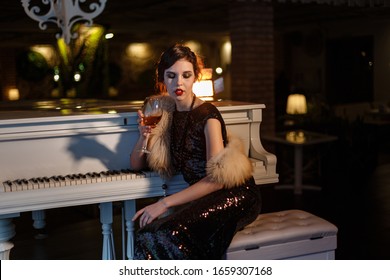 Portrait Of 20s Style Festive Beauty In A Restaurant. Young Beautiful Woman In Art-deco Style, In Black Dress In A Luxurious Interere Chicago 20s, The Era Of Gangsters. Singer  At The White Piano