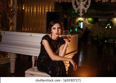 Portrait Of 20s Style Festive Beauty In A Restaurant. Young Beautiful Woman In Art-deco Style, In Black Dress In A Luxurious Interere Chicago 20s, The Era Of Gangsters. Singer  At The White Piano