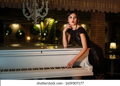 Portrait Of 20s Style Festive Beauty In A Restaurant. Young Beautiful Woman In Art-deco Style, In Black Dress In A Luxurious Interere Chicago 20s, The Era Of Gangsters. Singer  At The White Piano