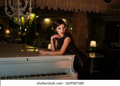 Portrait Of 20s Style Festive Beauty In A Restaurant. Young Beautiful Woman In Art-deco Style, In Black Dress In A Luxurious Interere Chicago 20s, The Era Of Gangsters. Singer  At The White Piano