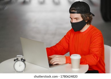 Portrait Of A 25-30 Year Old Man Wearing A Black Protective Mask, Cap And Red Sweatshirt. The Man Sits Alone At A Table In The Open Space, Drinking Coffee And Working On His Laptop. Freelance Concept