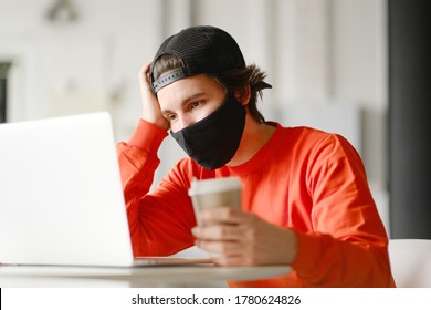 Portrait Of A 25-30 Year Old Man Wearing A Black Protective Mask, Cap And Red Sweatshirt. The Man Sits Alone At A Table In The Open Space, Drinking Coffee And Working On His Laptop. Freelance Concept