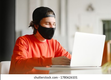 Portrait Of A 25-30 Year Old Man Wearing A Black Protective Mask, Cap And Red Sweatshirt. The Man Sits Alone At A Table In The Open Space, Drinking Coffee And Working On His Laptop. Freelance Concept