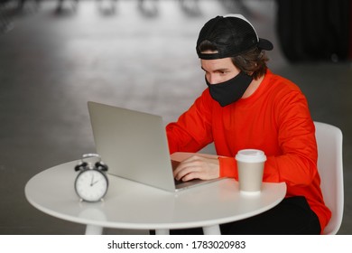 Portrait Of A 25-30 Year Old Man Wearing A Black Protective Mask, Cap And Red Sweatshirt. The Man Sits Alone At A Table In The Open Space, Drinking Coffee And Working On His Laptop. Freelance Concept