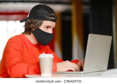 Portrait Of A 25-30 Year Old Man Wearing A Black Protective Mask, Cap And Red Sweatshirt. The Man Sits Alone At A Table In The Open Space, Drinking Coffee And Working On His Laptop. Freelance Concept