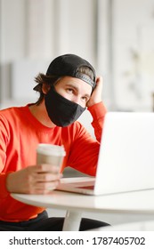 Portrait Of A 25-30 Year Old Man Wearing A Black Protective Mask, Cap And Red Sweatshirt. The Man Sits Alone At A Table In The Open Space, Drinking Coffee And Working On His Laptop. Freelance Concept