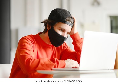 Portrait Of A 25-30 Year Old Man Wearing A Black Protective Mask, Cap And Red Sweatshirt. The Man Sits Alone At A Table In The Open Space, Drinking Coffee And Working On His Laptop. Freelance Concept