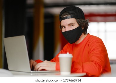 Portrait Of A 25-30 Year Old Man Wearing A Black Protective Mask, Cap And Red Sweatshirt. The Man Sits Alone At A Table In The Open Space, Drinking Coffee And Working On His Laptop. Freelance Concept