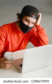 Portrait Of A 25-30 Year Old Man Wearing A Black Protective Mask, Cap And Red Sweatshirt. The Man Sits Alone At A Table In The Open Space, Drinking Coffee And Working On His Laptop. Freelance Concept