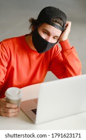 Portrait Of A 25-30 Year Old Man Wearing A Black Protective Mask, Cap And Red Sweatshirt. The Man Sits Alone At A Table In The Open Space, Drinking Coffee And Working On His Laptop. Freelance Concept