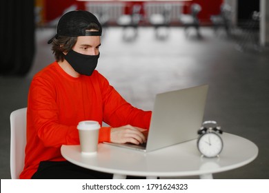 Portrait Of A 25-30 Year Old Man Wearing A Black Protective Mask, Cap And Red Sweatshirt. The Man Sits Alone At A Table In The Open Space, Drinking Coffee And Working On His Laptop. Freelance Concept