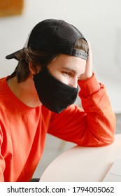 Portrait Of A 25-30 Year Old Man Wearing A Black Protective Mask, Cap And Red Sweatshirt. The Man Sits Alone At A Table In The Open Space, Drinking Coffee And Working On His Laptop. Freelance Concept