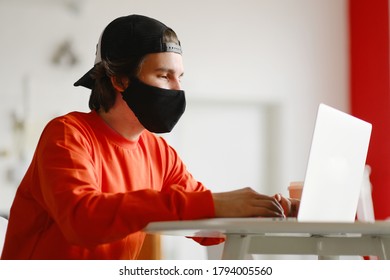 Portrait Of A 25-30 Year Old Man Wearing A Black Protective Mask, Cap And Red Sweatshirt. The Man Sits Alone At A Table In The Open Space, Drinking Coffee And Working On His Laptop. Freelance Concept