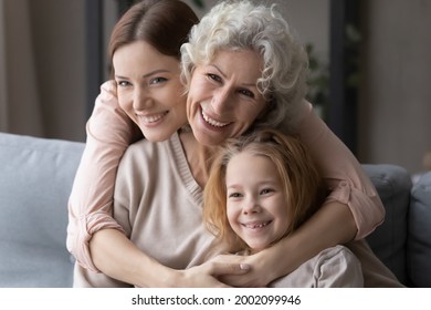 Portrait of 3 generations family. Happy young female embrace from back smiling mature mother and preteen child girl. Caring grey haired grandma sit on sofa cuddle little grandkid and adult daughter - Powered by Shutterstock