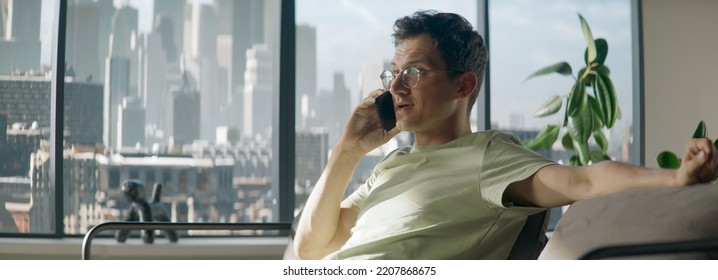 Portrait Of 30s Caucasian Male Having A Phone Call At Home. Sitting On A Sofa In A Modern Apartment, Living Room, Big Windows, Urban Background. Daytime Shot