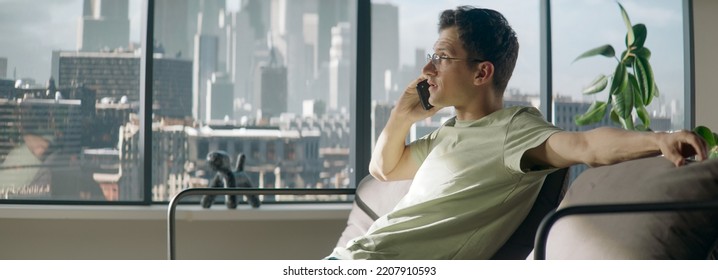 Portrait Of 30s Caucasian Male Having A Phone Call At Home. Sitting On A Sofa In A Modern Apartment, Living Room, Big Windows, Urban Background. Daytime Shot