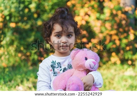 A portrait of a 3-year-old Brazilian girl with curly hair, outside in a public park, holding her pink teddy bear. The child is looking at the camera but not smiling