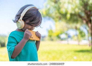 Portrait Of 4 Years Old Boy Using Headphones And Smartphone. Child Using A Tablet And Listening To Music In The Park - Powered by Shutterstock