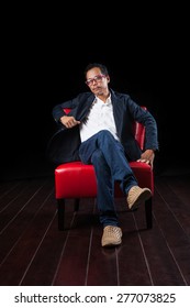 Portrait Of 45 Years Old Asian Man Sitting On Red Sofa Against Black Background 