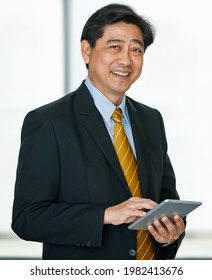 Portrait 50s Asian Male Executive Visionary Business Male Owner Wearing Formal Suit And Necktie, Standing Indoor Office, Using Technology Tablet, Touching The Screen, Smiling Confidently And Reliably