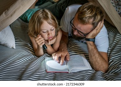 Portrait of a 6 year old boy and his father reading a book in teepee tent. Father and son with flashlight reading book under blanket at home.  - Powered by Shutterstock