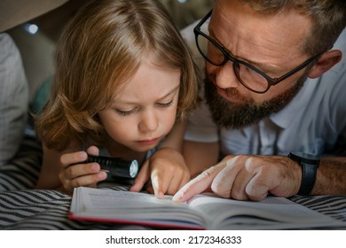 Portrait of a 6 year old boy and his father reading a book in teepee tent. Father and son with flashlight reading book under blanket at home. 