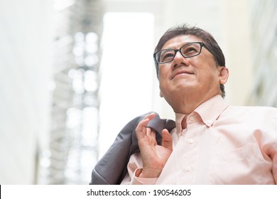 Portrait Of 60s Asian Chinese CEO Boss Smiling And Looking Away. Senior Male Businessman, Real Modern Office Building As Background.