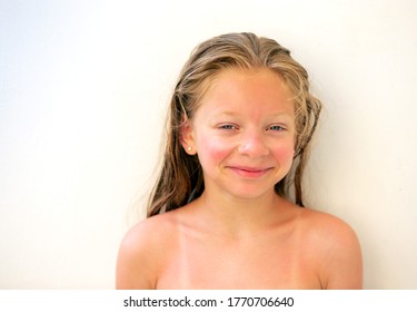 Portrait Of A 9-year-old Girl With Long Wet Blond Hair And Blue Eyes, With A Sunburn On Her Skin And The Trace Of A Swimsuit, Smiling Cheerfully And Looking At The Frame, On A Light Background