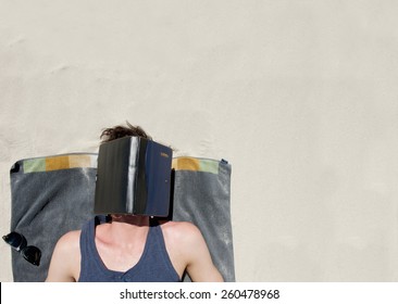 Portrait From Above Of Young Man Lying Down On A Towel At The Beach With Book Covering Face