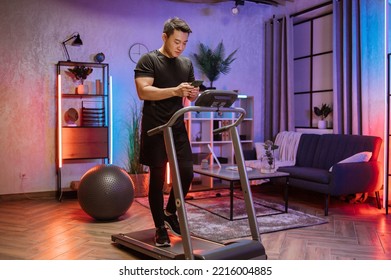 Portrait of active asian man in sportswear using smartphone, running using treadmill at home at night. Coronavirus Covid 19 social distance. Home workout, sports, stay at home, remote leisure concept. - Powered by Shutterstock