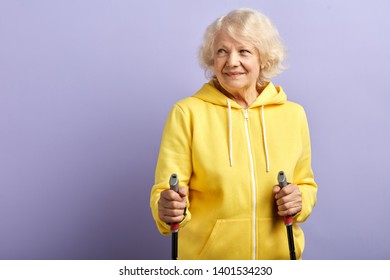 Portrait of active elderly female retiree wearing yellow sportive outfit practicing Nordic walking with poles, posing isolated in studio with purple background. - Powered by Shutterstock