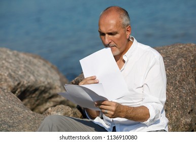 Portrait of an active and energetic mature european business man in a white shirt,working with documents and letters during a summer holiday on a rocky beach on the sea.concept of life after 50 years. - Powered by Shutterstock