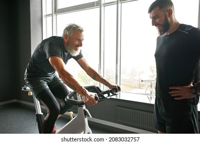 Portrait Of Active Mature Man With Gray Hair And Beard, Working Out On Gym Bike Next To Young Personal Trainer. Endurance Training While Retirement. Personal Resilience Concept.