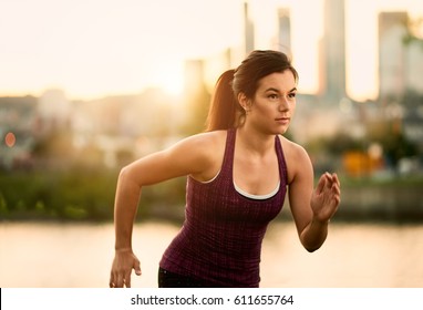 Portrait Of Active Millenial Woman Jogging At Dusk With An Urban Cityscape And Sunset In The Background