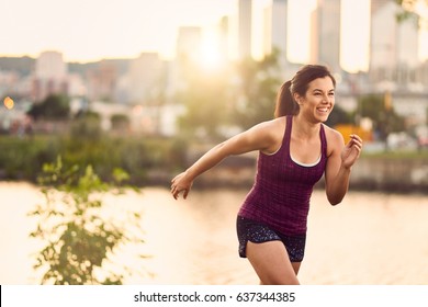 Portrait Of Active Millenial Woman Jogging At Dusk With An Urban Cityscape And Sunset In The Background