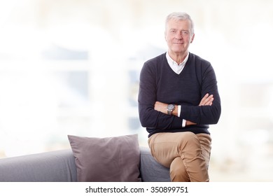 Portrait Of Active Senior Man Sitting At Sofa With Arms Crossed While Looking At Camera And Smiling.