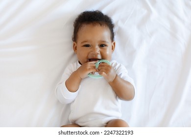 Portrait Of Adorable African American Baby Biting Teether And Looking At Camera While Lying On Bed At Home, Cute Black Infant Child Laughing And Playing With Toy While Relaxing In Bedroom, Top View