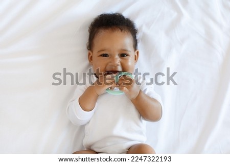 Portrait Of Adorable African American Baby Biting Teether And Looking At Camera While Lying On Bed At Home, Cute Black Infant Child Laughing And Playing With Toy While Relaxing In Bedroom, Top View