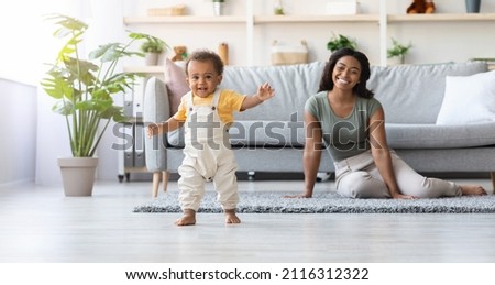 Portrait Of Adorable African American Infant Boy Making His First Steps At Home, Cute Little Black Toddler Child Wearing Jumpsuit Walking In Living Room, His Happy Mom Smiling On Background, Panorama